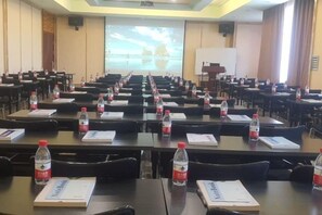Une salle de conférence avec des rangées de tables, des chaises, des plaques nominatives, des bouteilles d’eau et un écran de projection.