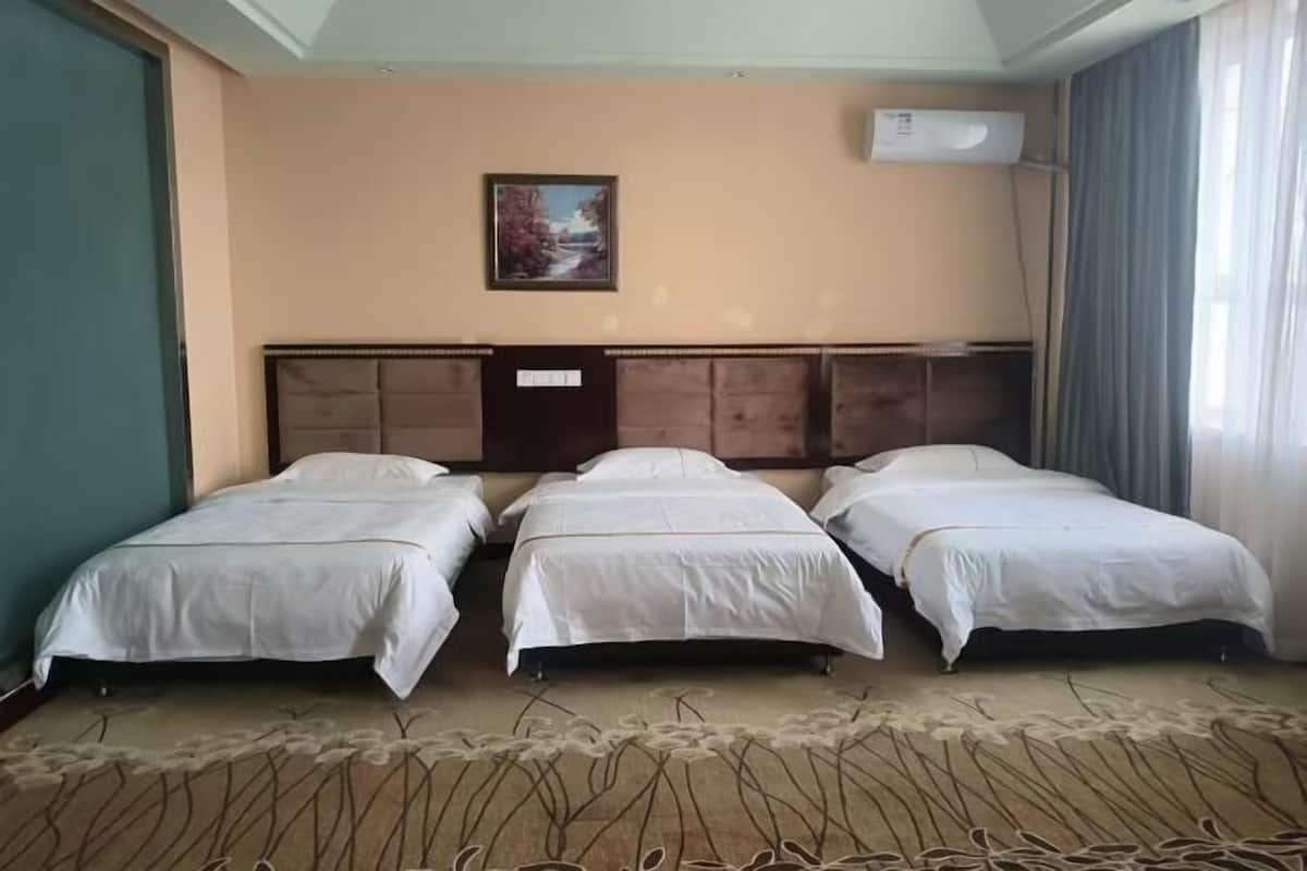 A hotel room with three single beds, a large headboard, a painting on the wall, and an air conditioning unit.