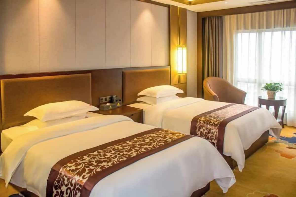 A hotel room with two beds, a nightstand, a chair, a lamp, and a window with curtains.