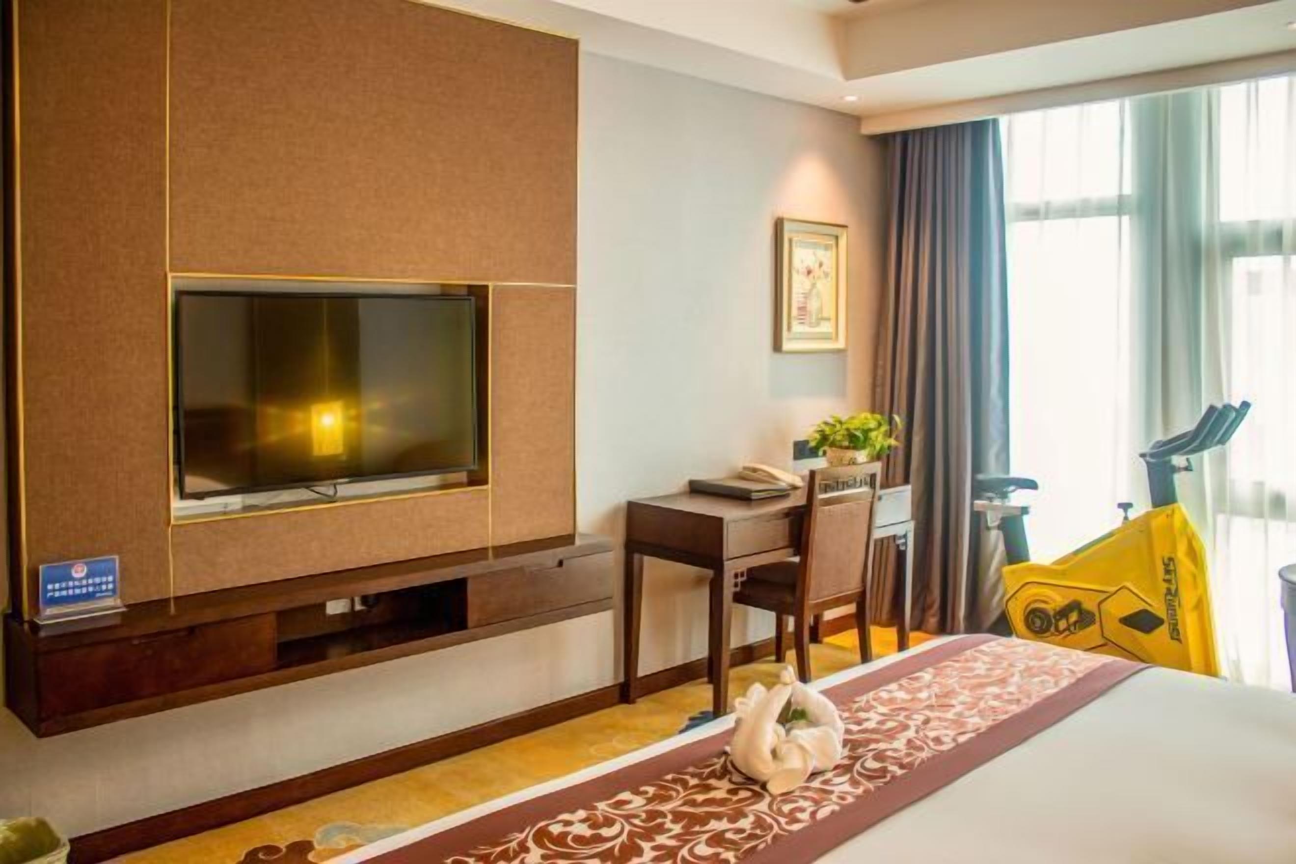 Habitación de hotel con una gran televisión de pantalla plana montada en la pared, un escritorio con una silla, una cama con un corredor decorativo y una ventana con cortinas.