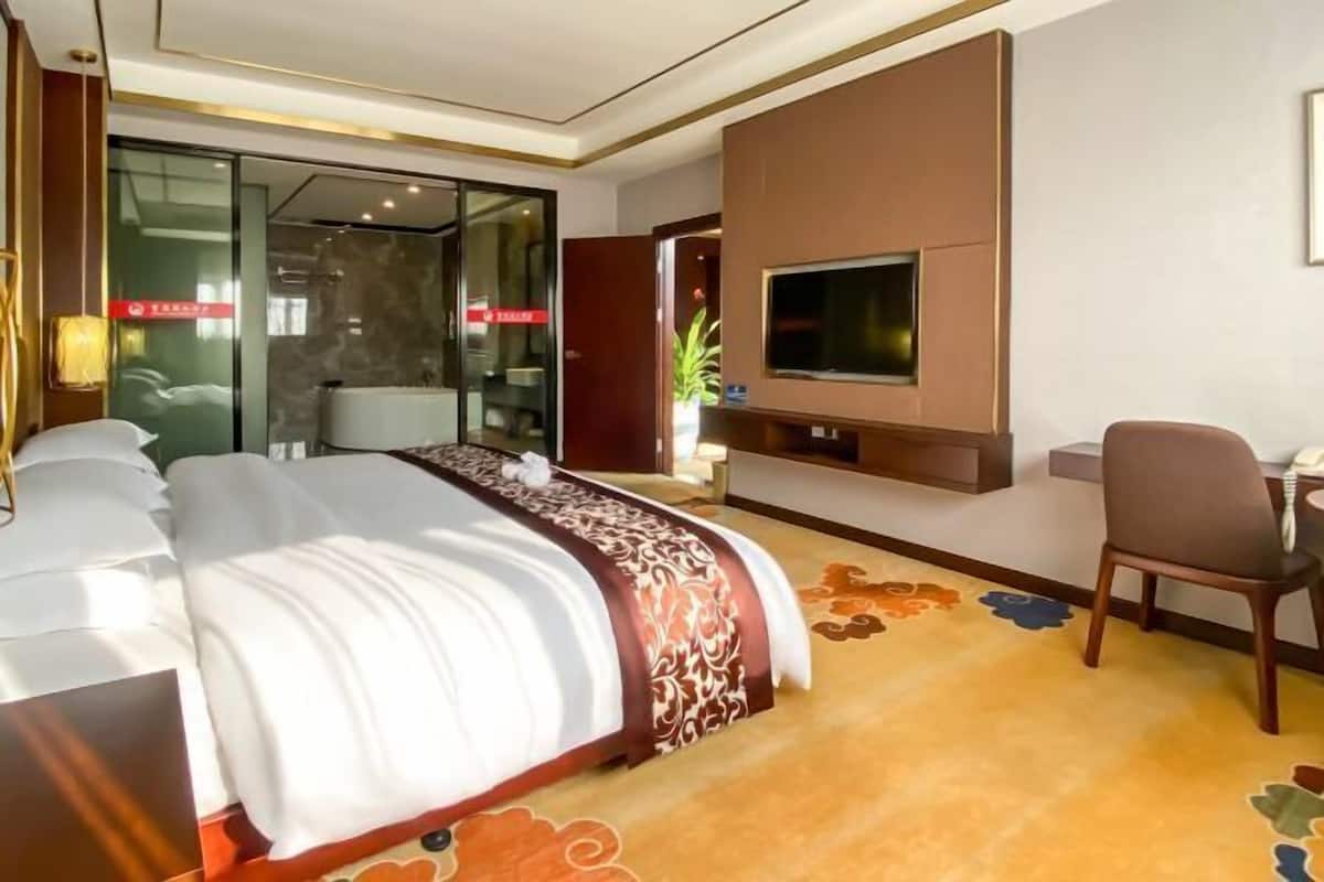 A hotel room with a large bed, a desk with a chair, a television mounted on the wall, and a glass door leading to a bathroom.