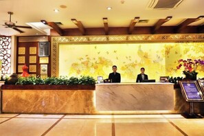 Ein Hotel-Empfangsbereich mit Marmor-Theke, zwei Rezeptionisten, einer Wand mit floralen Verzierungen und einem Deckenventilator.