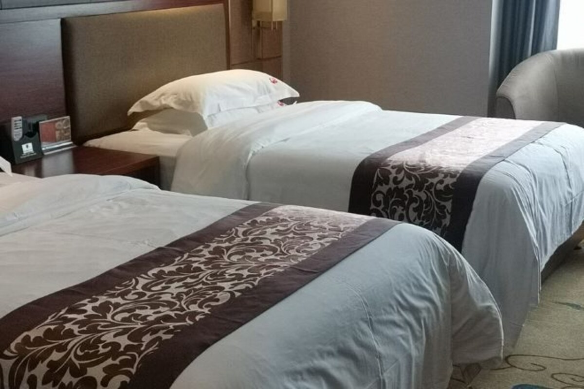 A hotel room with two beds, each with a white bedspread and a brown decorative trim. There is a nightstand with a lamp and a small table with a clock.