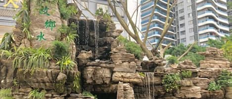 A rock formation with a waterfall and greenery, surrounded by high-rise buildings.