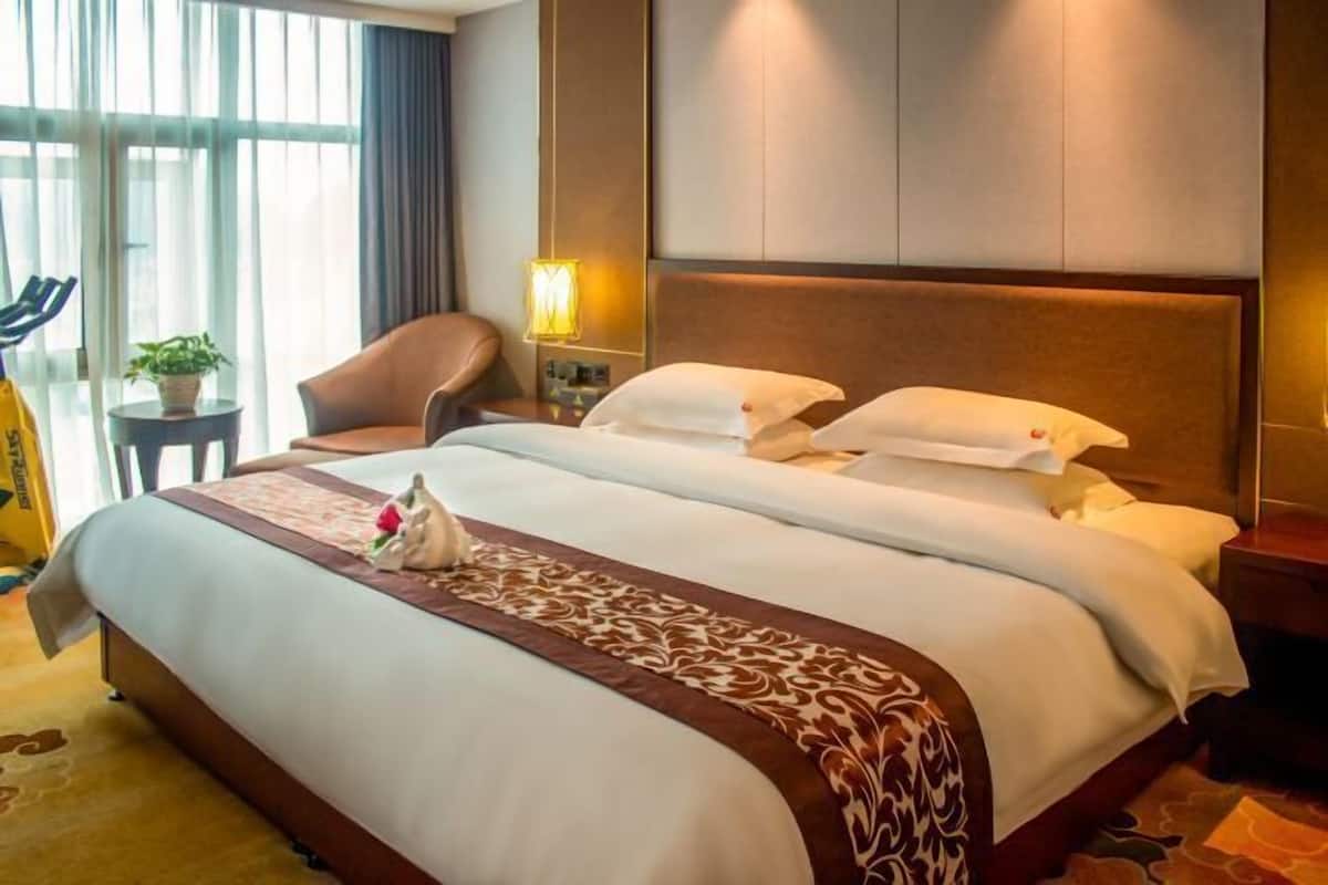 A hotel room with a large bed, a chair, a small table with a lamp, and a view of the outdoors through large windows.