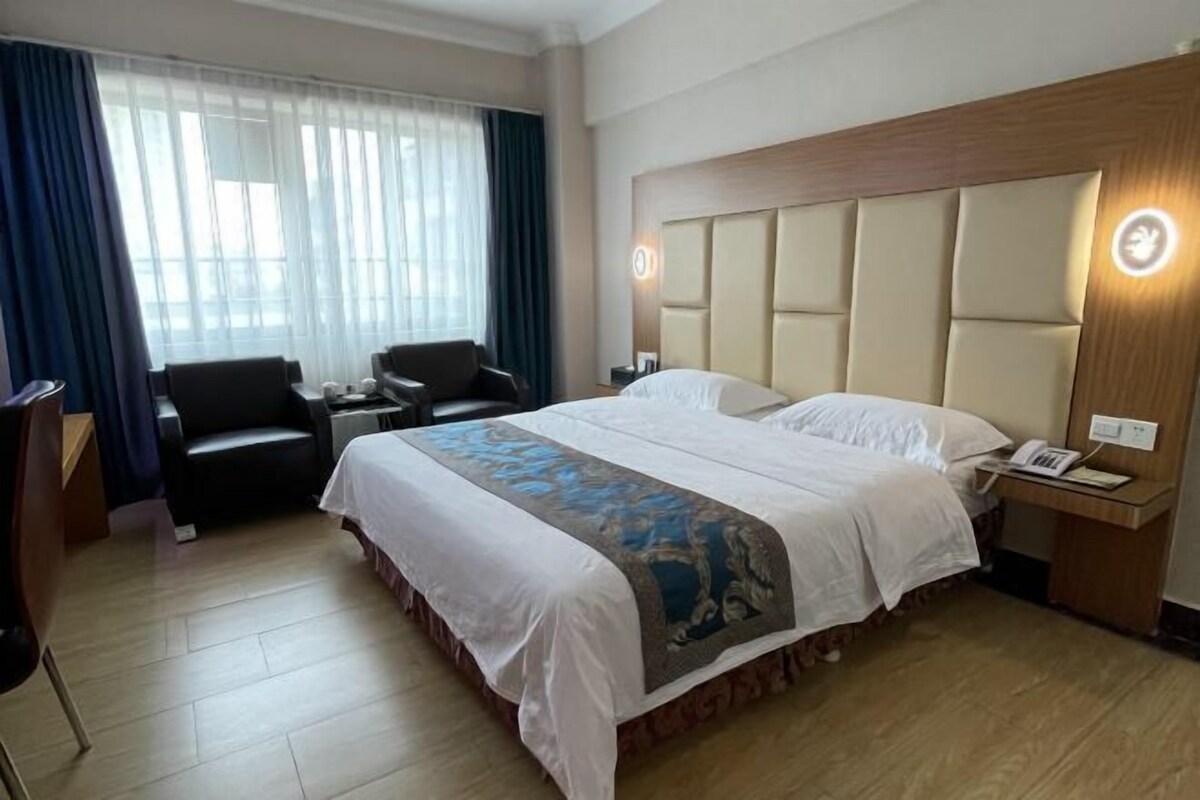 A hotel room with a large bed, a desk with a telephone, a chair, and a window with curtains.