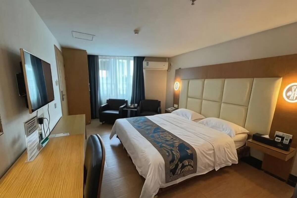 A hotel room with a bed, a desk, a chair, a TV, a bedside table, and a window with curtains.
