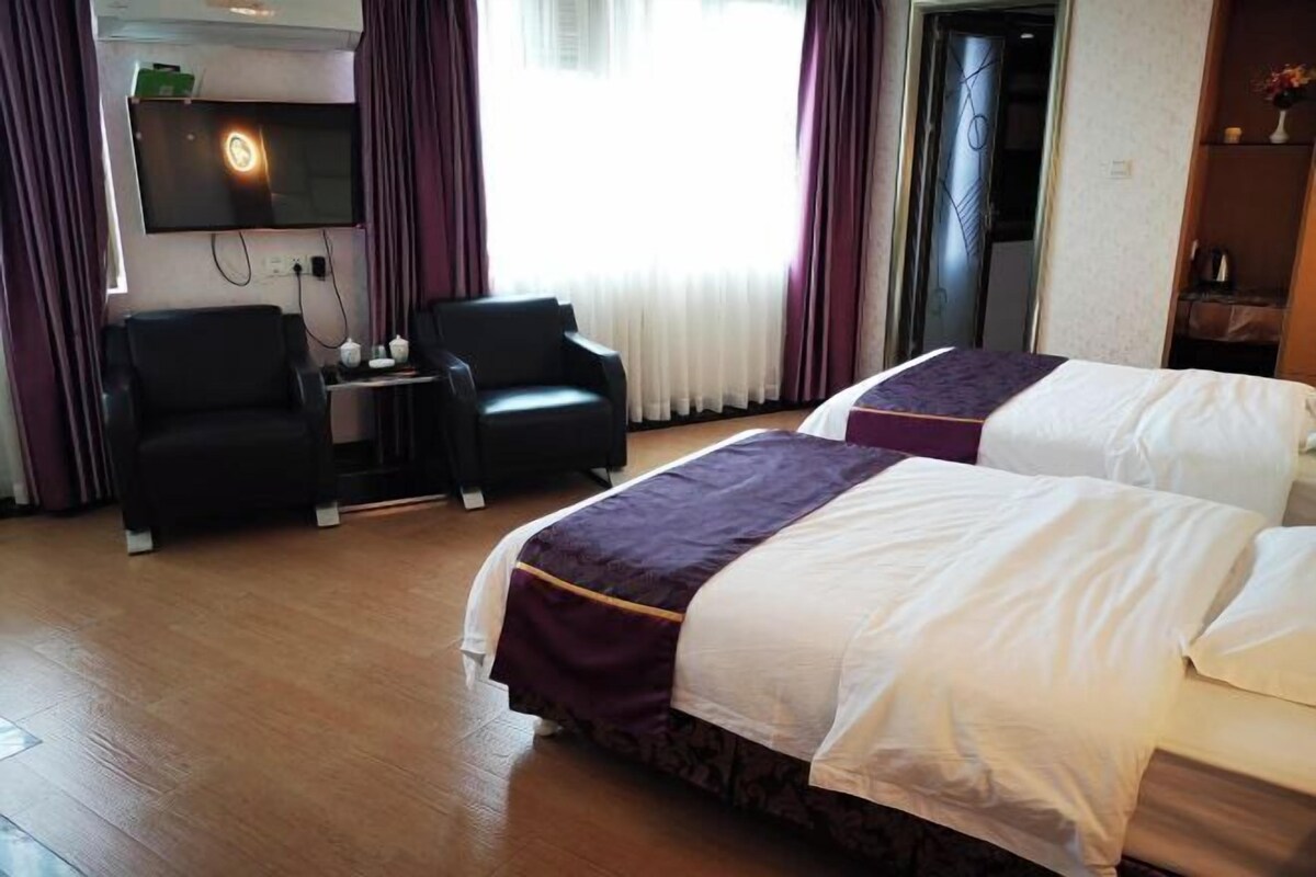 A hotel room with two beds, a TV mounted on the wall, a small table with a chair, and a window with curtains.