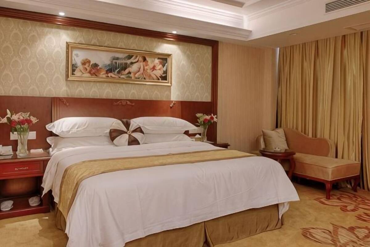 A hotel room with a large bed, a nightstand, a chair, a painting on the wall, and a window with curtains.
