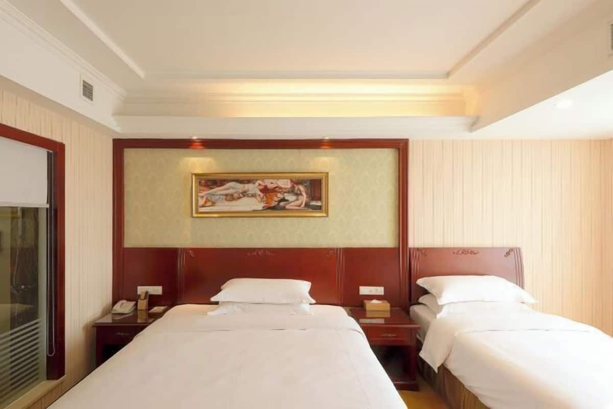 A hotel room with two single beds, a painting on the wall, and a window with blinds.