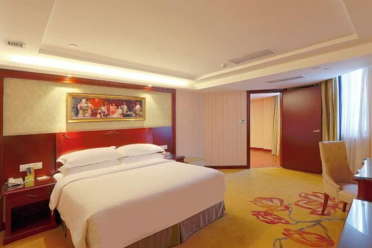 A hotel room with a large bed, a painting on the wall, a desk, and a chair.
