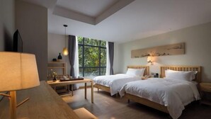 Ein Hotelzimmer mit zwei Betten, einem Schreibtisch, einer Lampe, einem Fenster mit Blick auf Grünflächen und einem Wandbild.