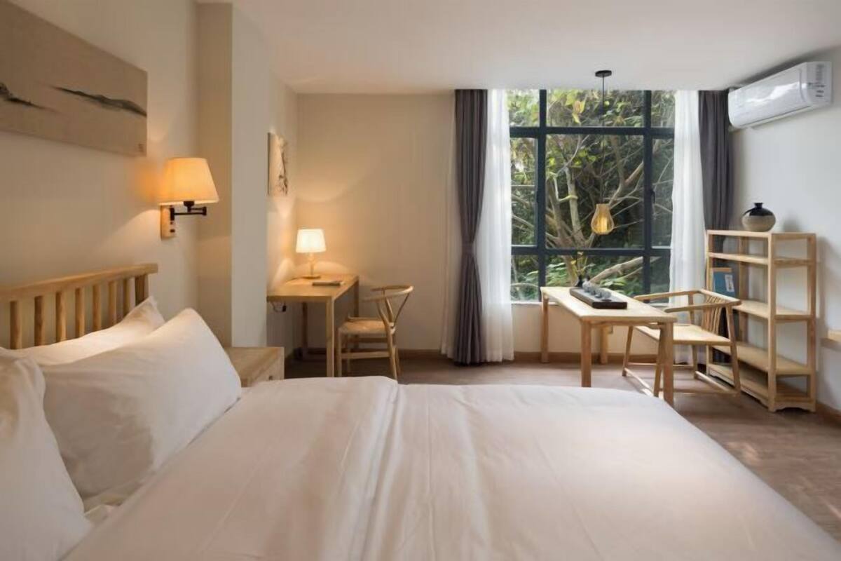 Een hotelkamer met een bed, bureau, stoel, lamp, raam met uitzicht op groen en een plank met een vaas.