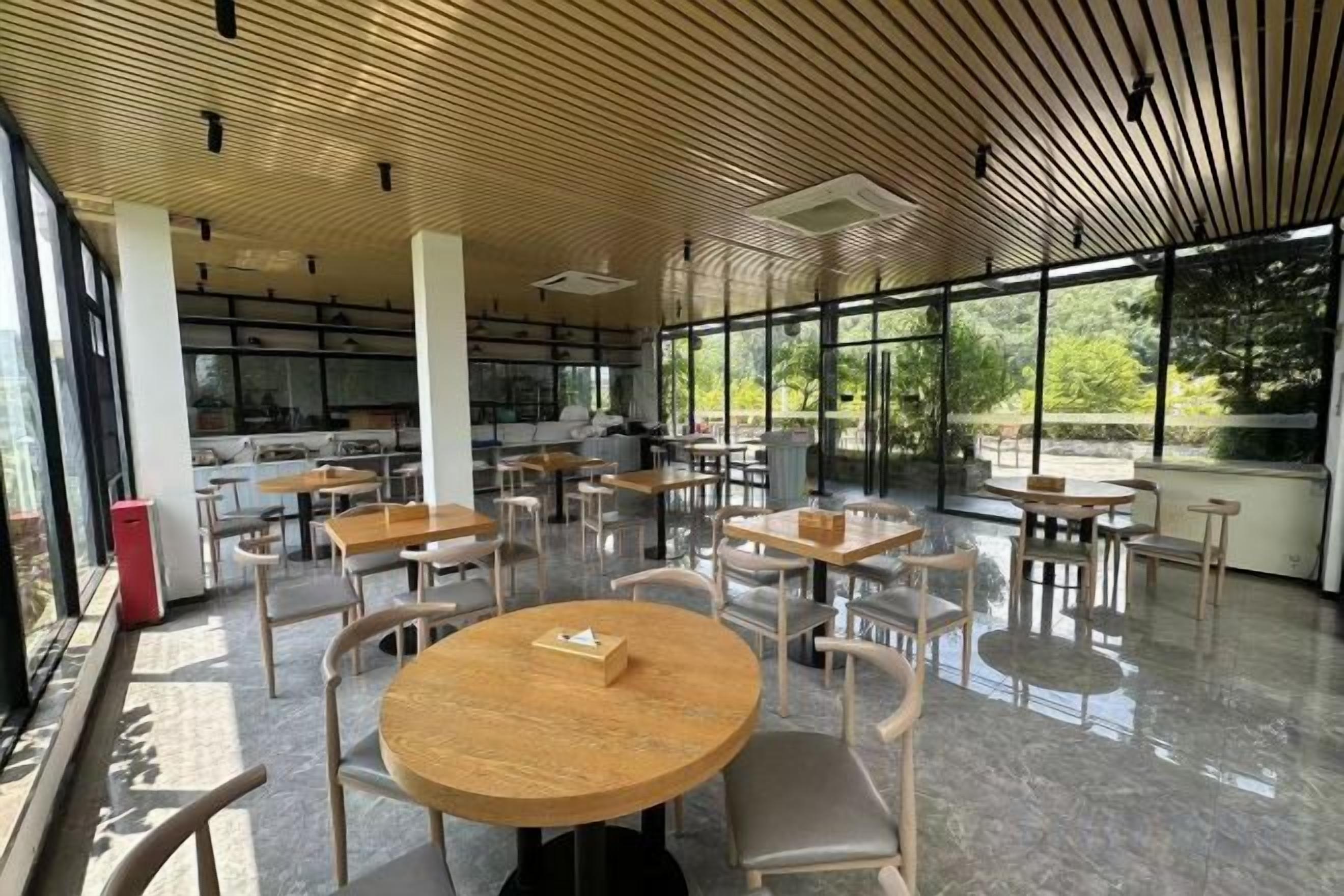 A modern indoor space with round tables and chairs, large windows, and a wooden ceiling.