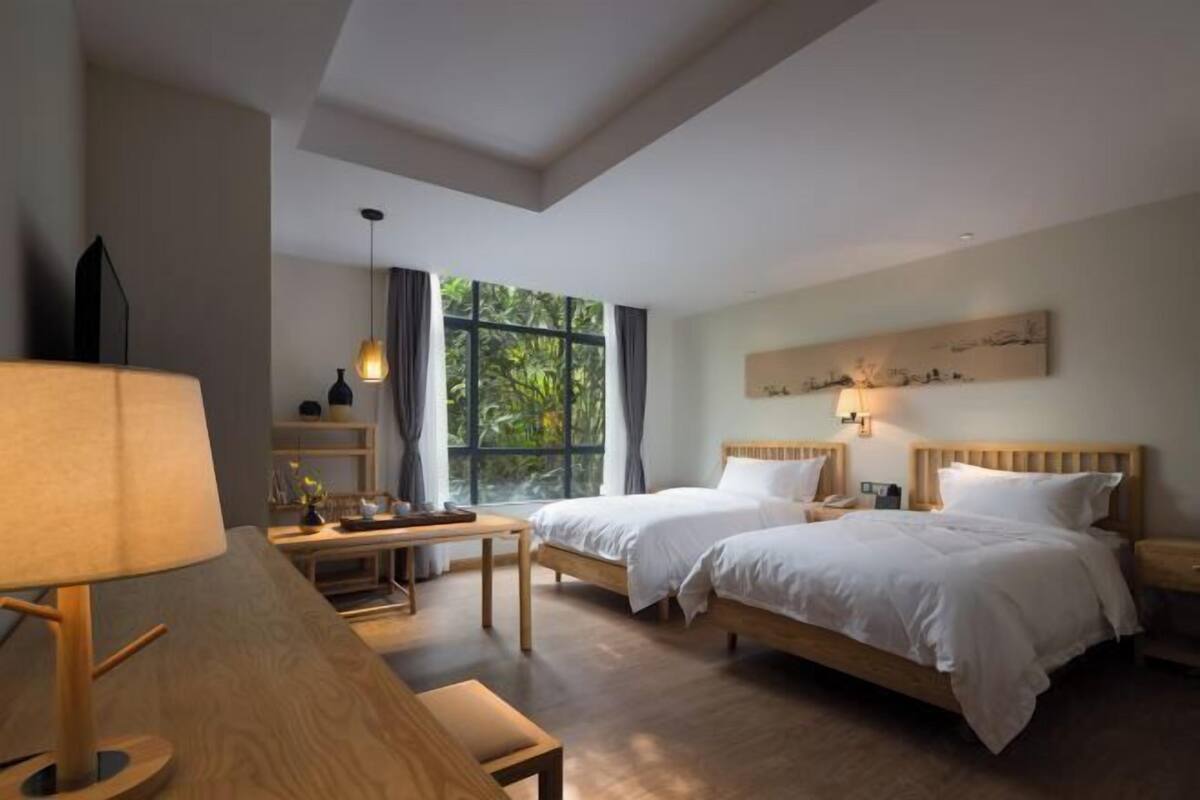 Een hotelkamer met twee bedden, een bureau, een lamp en een groot raam met uitzicht op groen.