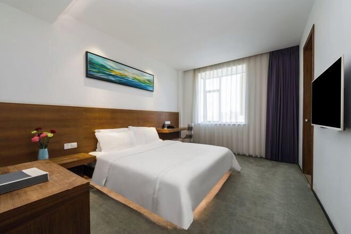 Una habitación de hotel con una cama, una mesita de noche, un televisor, una ventana con cortinas y un cuadro en la pared.
