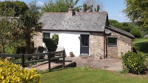 Un pequeño cottage de piedra con fachada blanca, tejado gris y un jardín con cerca de madera y plantas en macetas.