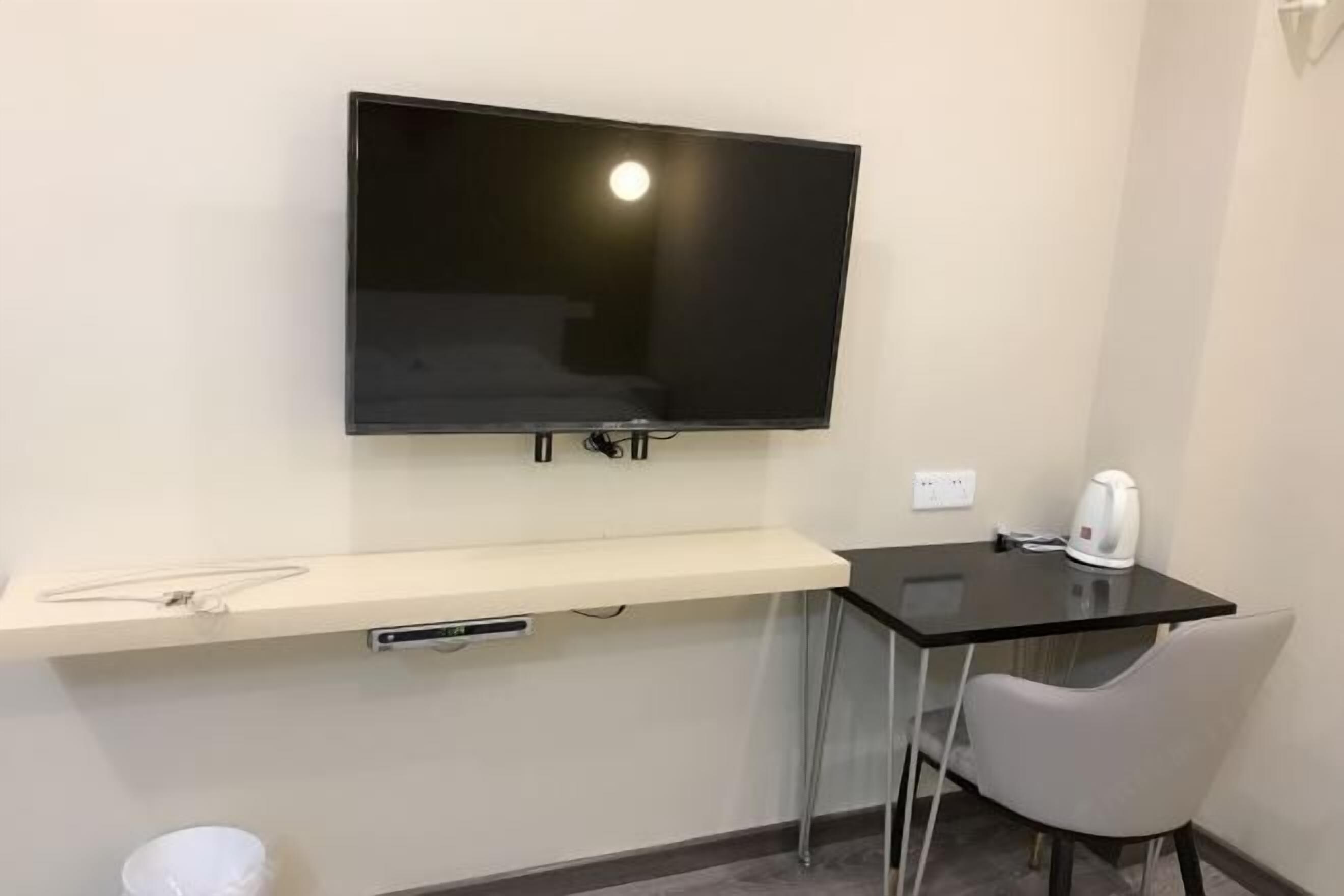 A small room with a flat-screen TV mounted on the wall, a small table with a kettle, and a chair.