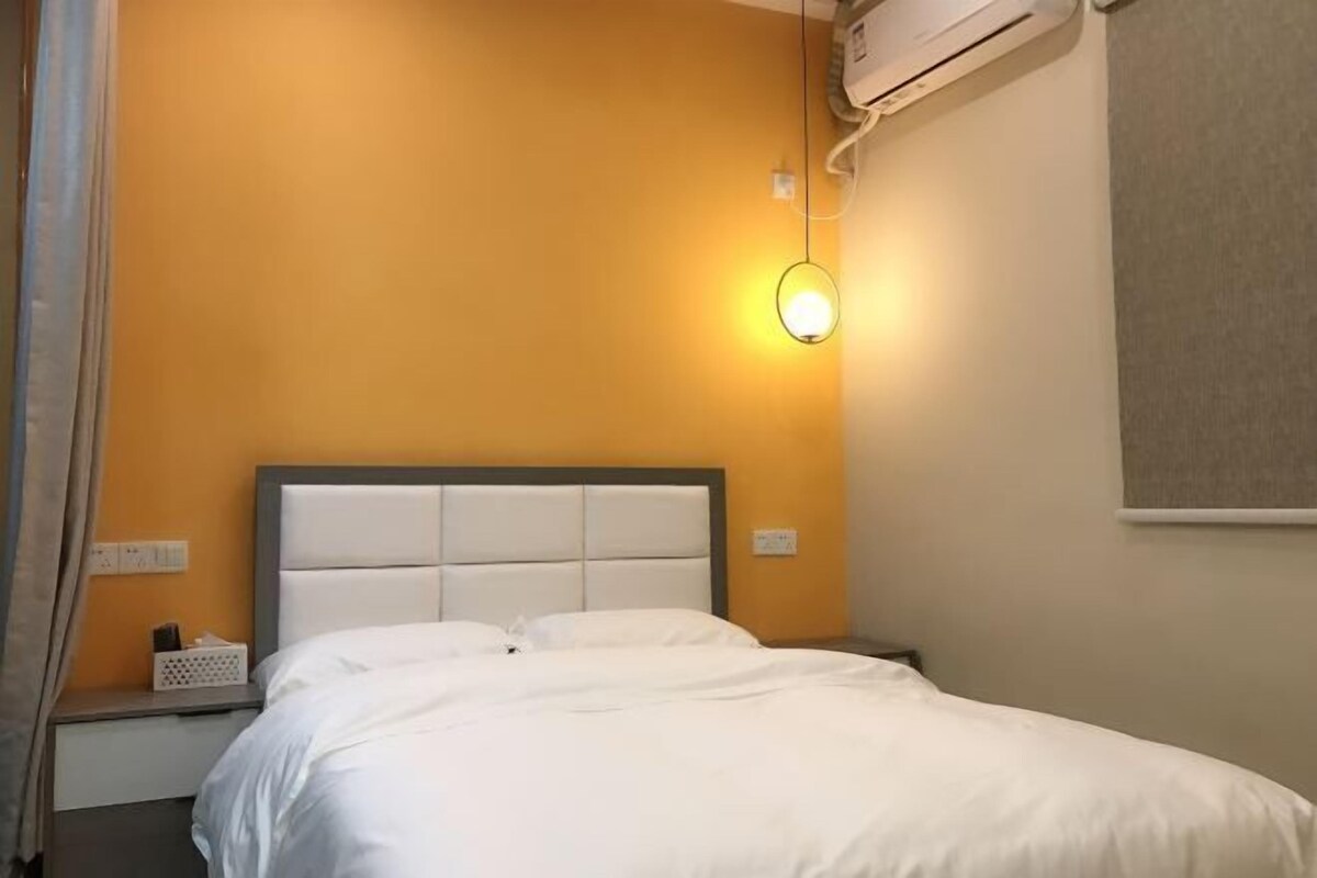 A hotel room with a bed, a nightstand, an air conditioning unit, and a wall-mounted light fixture.