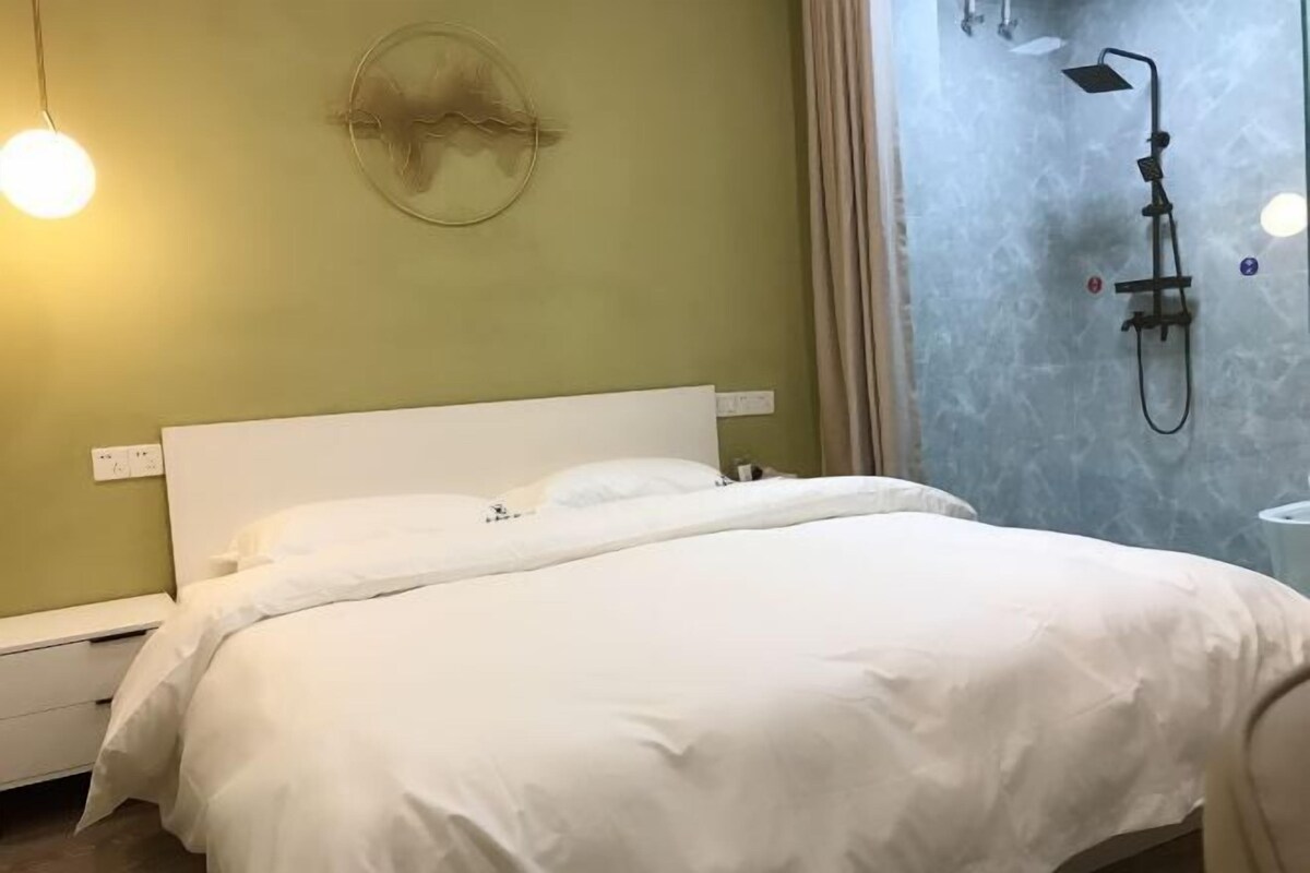 A neatly made bed with white bedding in a room with a green wall, a round wall decoration, and a shower area with a glass door.