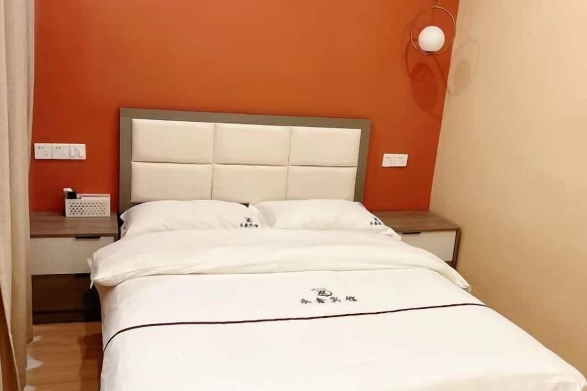 A neatly made bed with a white headboard and pillows, a bedside table with a lamp, and a wall socket.