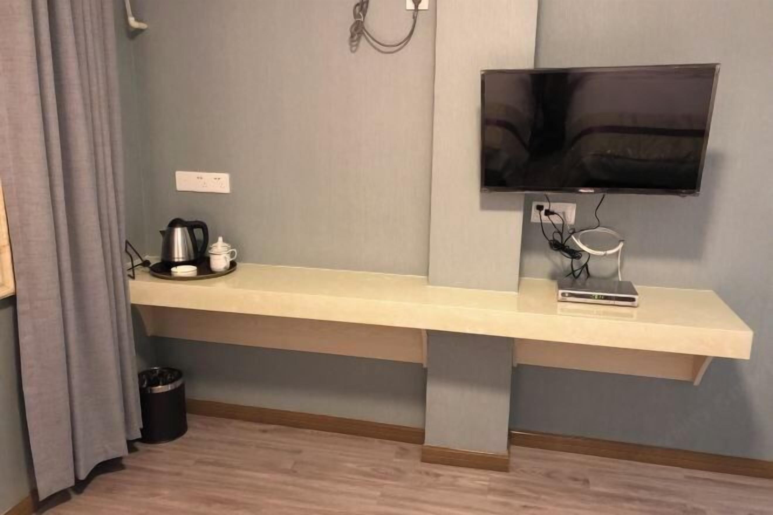 Ein modernes Hotelzimmer mit einer langen, hellen Bank an einer grauen Wand, einem flachbildfernseher darüber und einem kleinen Tisch mit Wasserkocher und Tassen.