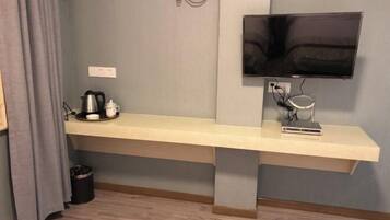 A modern hotel room with a long, light-colored bench against a grey wall, a flat-screen TV mounted above it, and a small table with a kettle and cups.