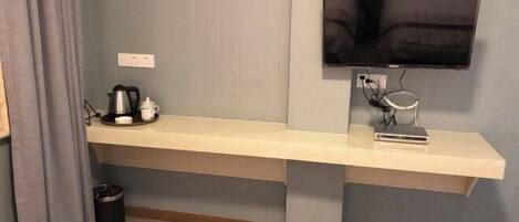 Ein modernes Hotelzimmer mit einer langen, hellen Bank an einer grauen Wand, einem flachbildfernseher darüber und einem kleinen Tisch mit Wasserkocher und Tassen.