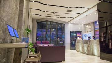 A modern hotel lobby with a reception desk, seating area, and glass walls offering views of the city.