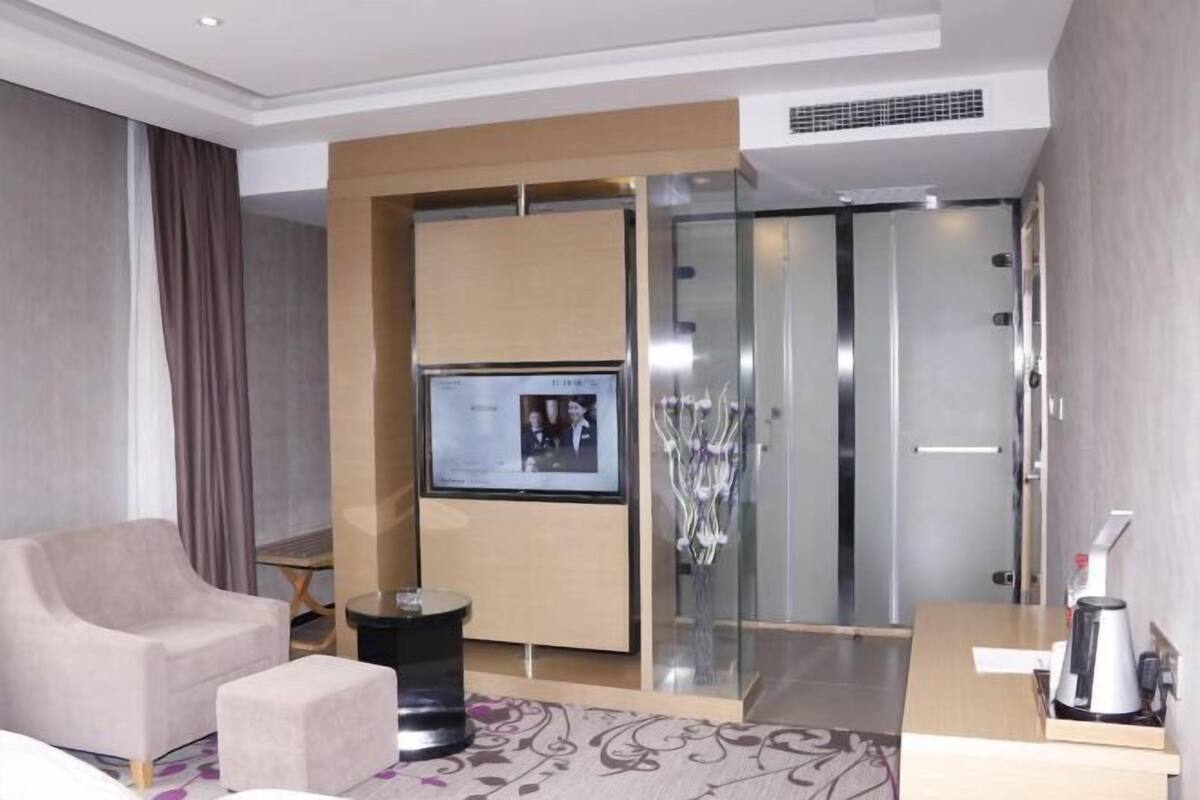Moderni hotellihuone, jossa on sohva, pieni pöytä, televisio, lasiseinäinen suihkutila ja kahvinkeitin.