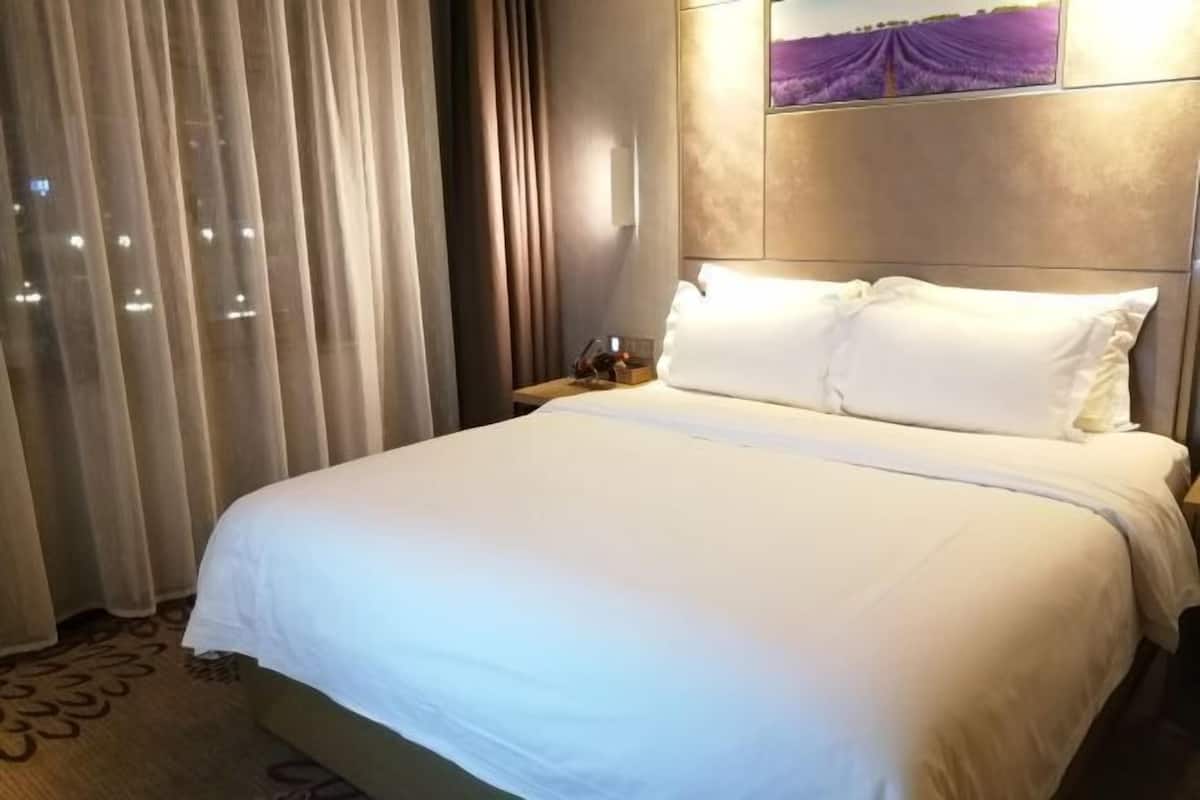 Hotellihuone, jossa on suuri sänky, verhot, yöpöytä ja seinälle ripustettu taideteos.