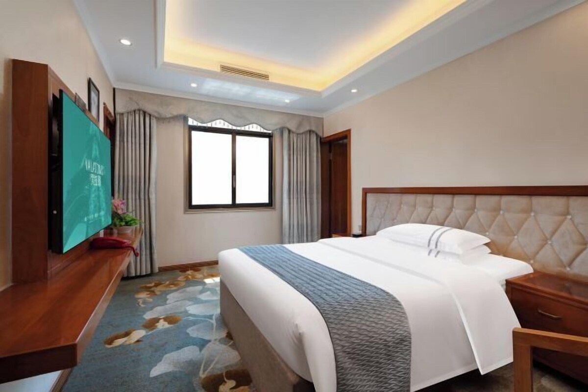 A hotel room with a large bed, a flat-screen TV, a wooden nightstand, and a window with curtains.