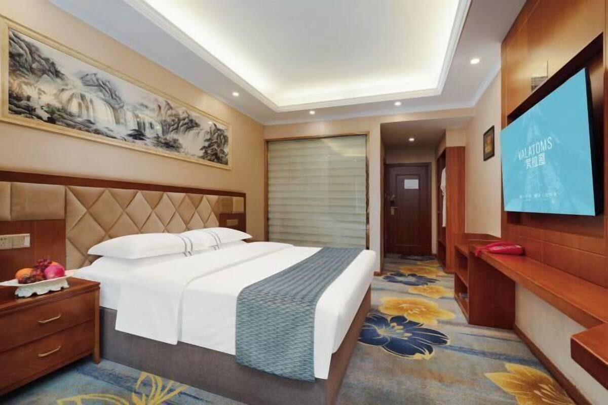 Une chambre d’hôtel avec un lit, une table de chevet, une télévision, un tableau et une moquette à motifs floraux.