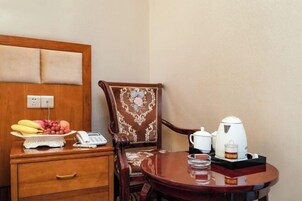 Una habitación de hotel con una mesita de noche de madera, un teléfono, un frutero, una silla con un cojín estampado, una mesa redonda con una tetera, un vaso y una bandeja.