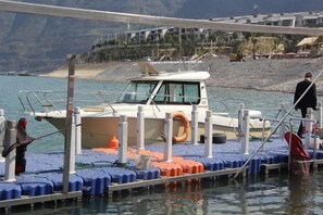 Un bateau était amarré à un quai, avec une bouée de sauvetage fixée à celui-ci. L’eau l’entourait, et un paysage montagneux se dressait à l’arrière-plan.