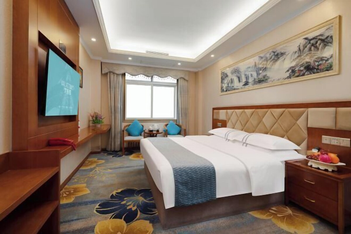 A hotel room with a large bed, a TV on the wall, a painting, and a desk with a chair.