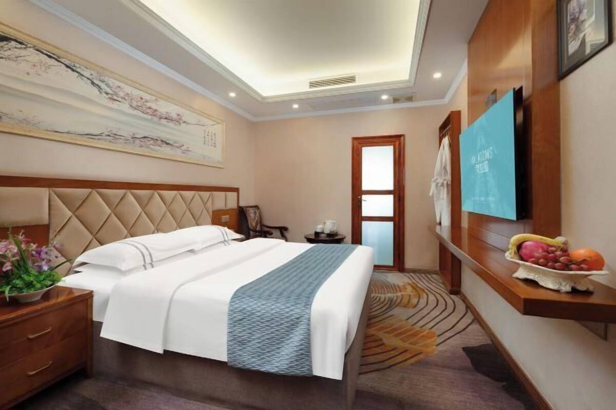Une chambre d’hôtel avec un lit, une table de chevet, un téléviseur fixé au mur, un panier de fruits et un grand tableau.