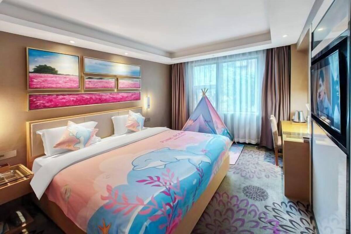 A hotel room with a bed, a TV, a window with curtains, and artwork on the wall.