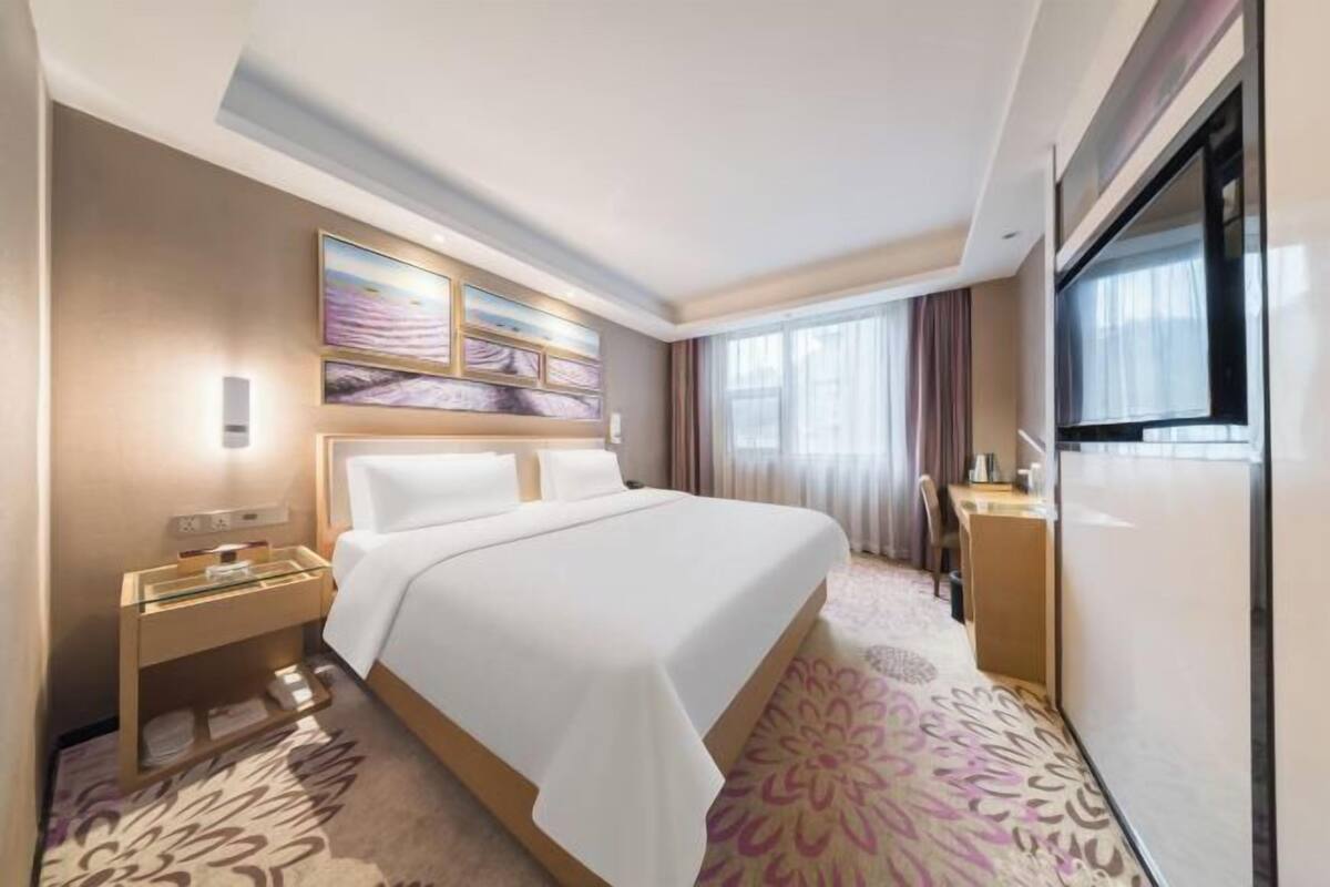 A hotel room with a bed, bedside table, artwork, curtains, and a television.
