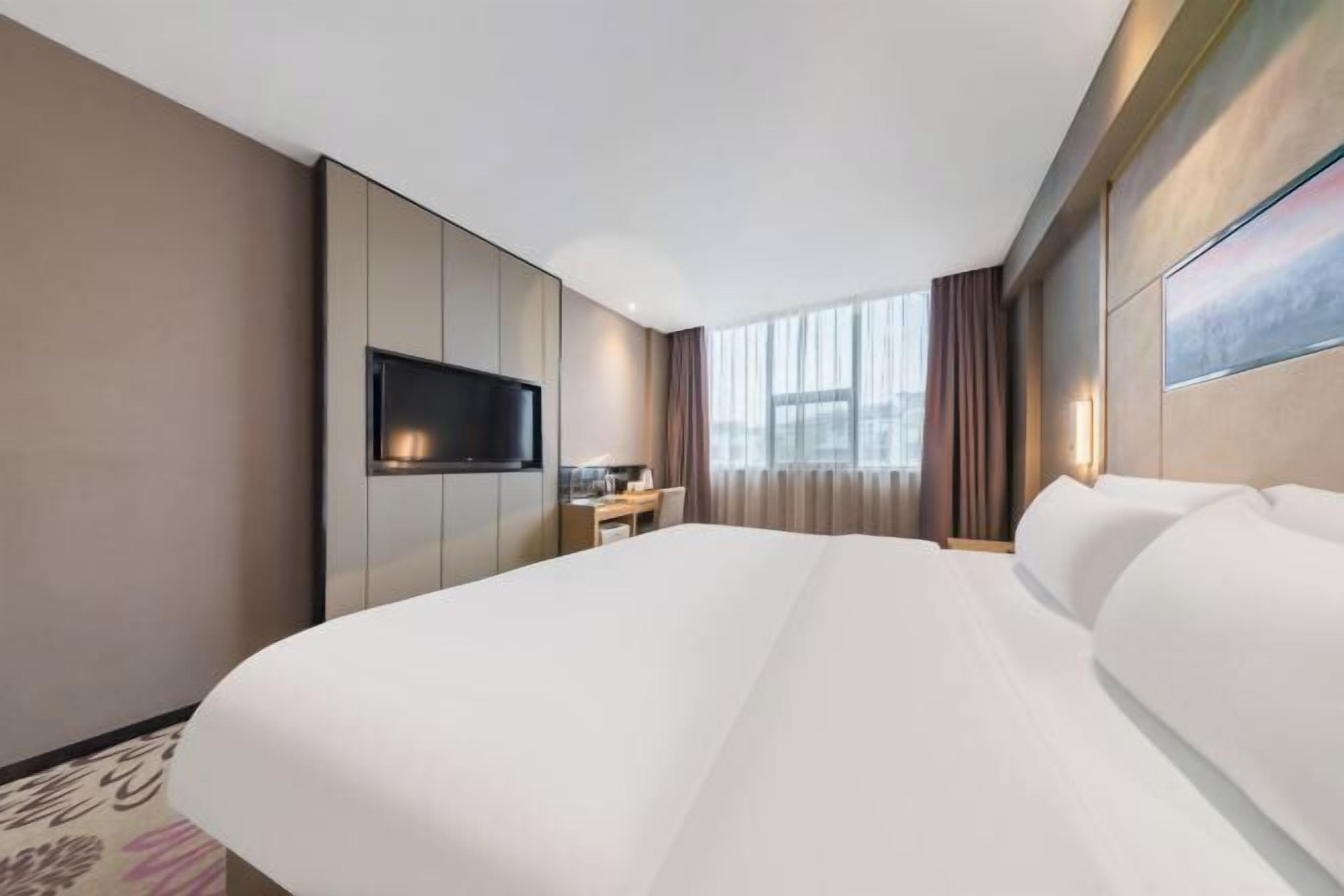 A hotel room with a large bed, a flat-screen TV mounted on the wall, a desk with a chair, and a window with curtains.