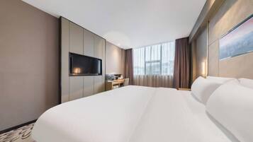 A hotel room with a large bed, a flat-screen TV mounted on the wall, a desk with a chair, and a window with curtains.