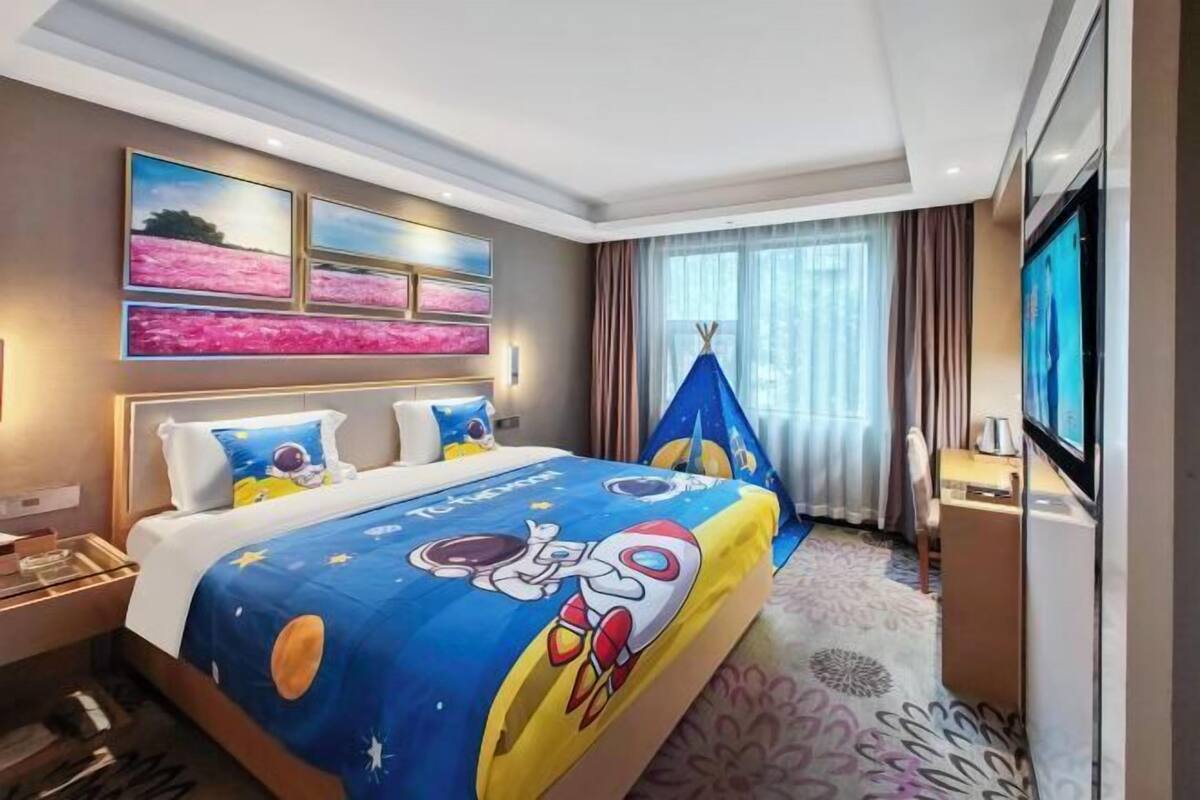 A hotel room with a bed, a TV, a desk, and a window with curtains.