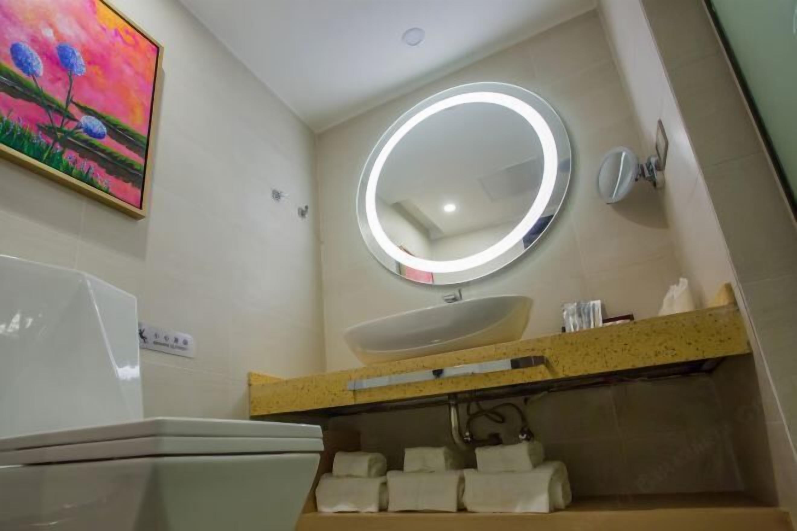 Une salle de bain avec un miroir rond, un lavabo et des toilettes. Il y a un tableau au mur et une étagère avec des serviettes.