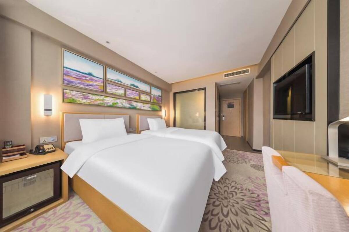 A hotel room with a bed, bedside table, lamp, artwork, and a television.