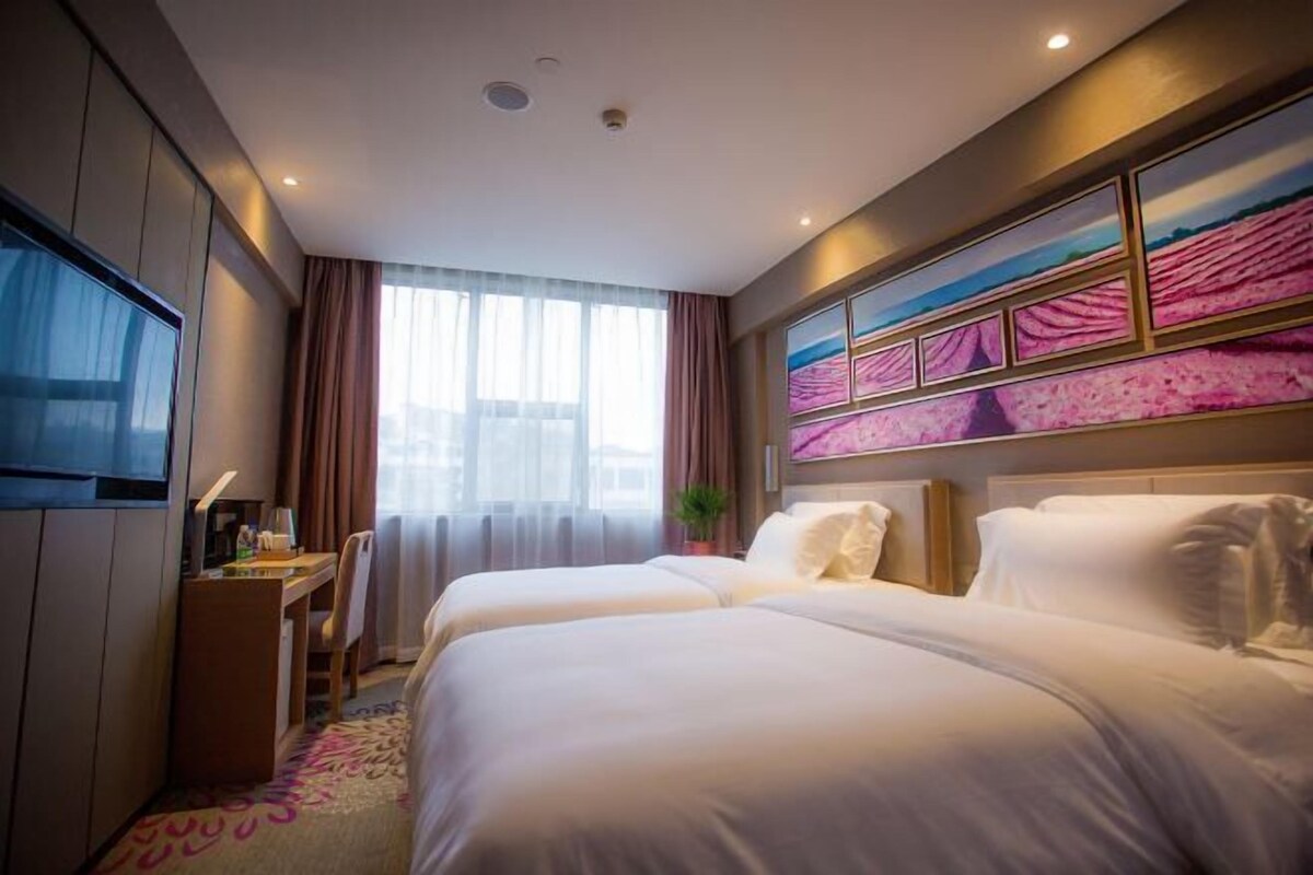 Quarto de hotel com duas camas, uma escrivaninha, televisão e uma janela grande com cortinas.