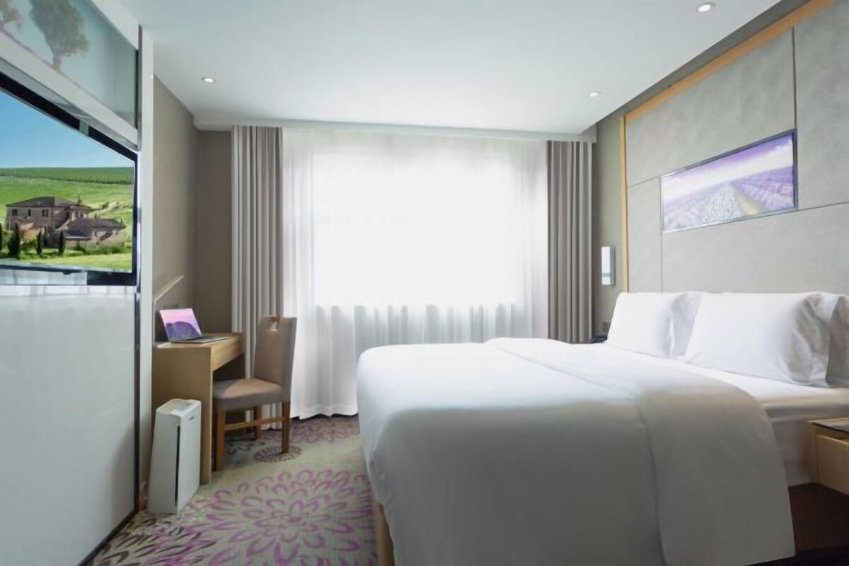 Quarto de hotel com uma cama grande, uma mesa com um laptop, uma cadeira e vista para uma casa pela janela.