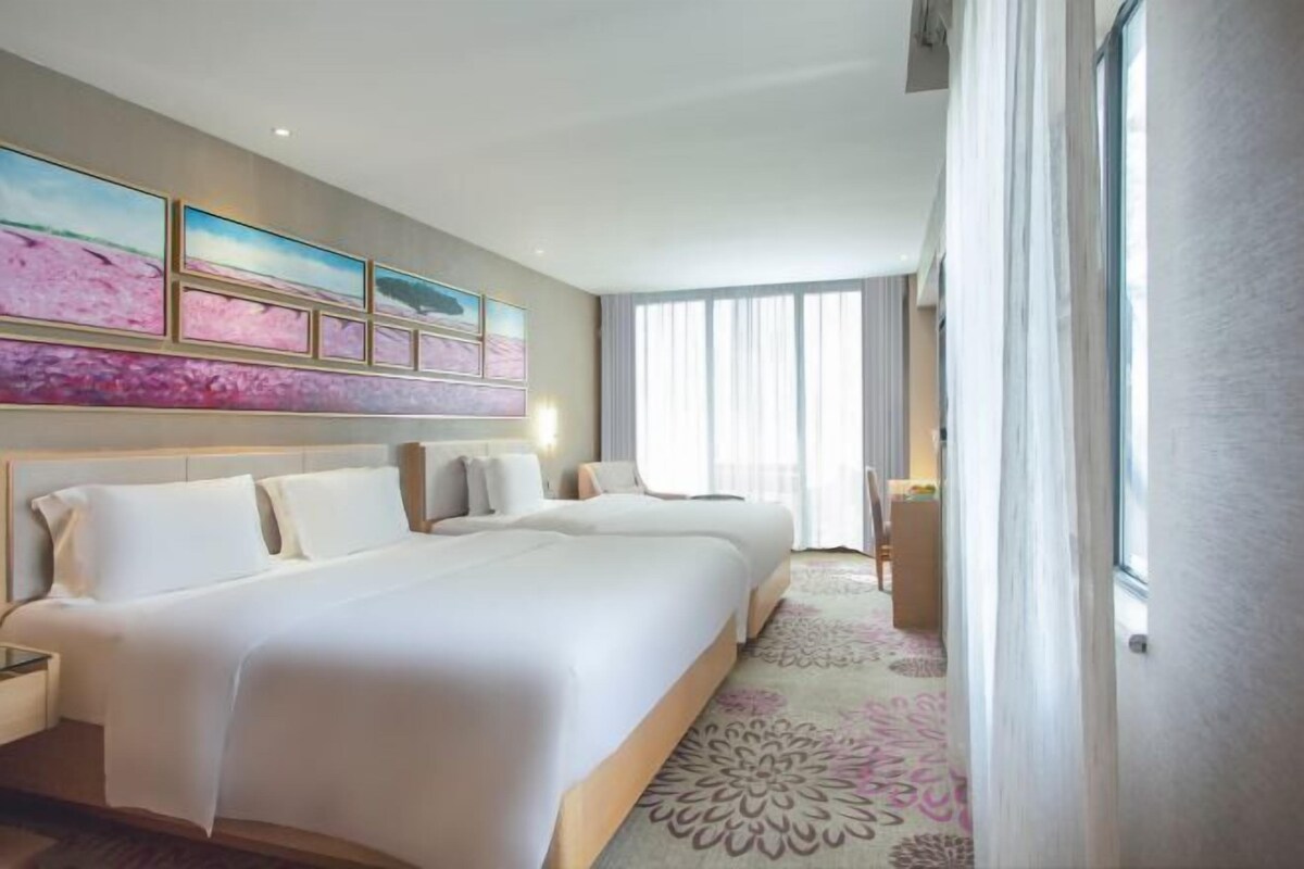 Quarto de hotel com duas camas, uma janela grande com cortinas e uma parede com obras de arte emolduradas.