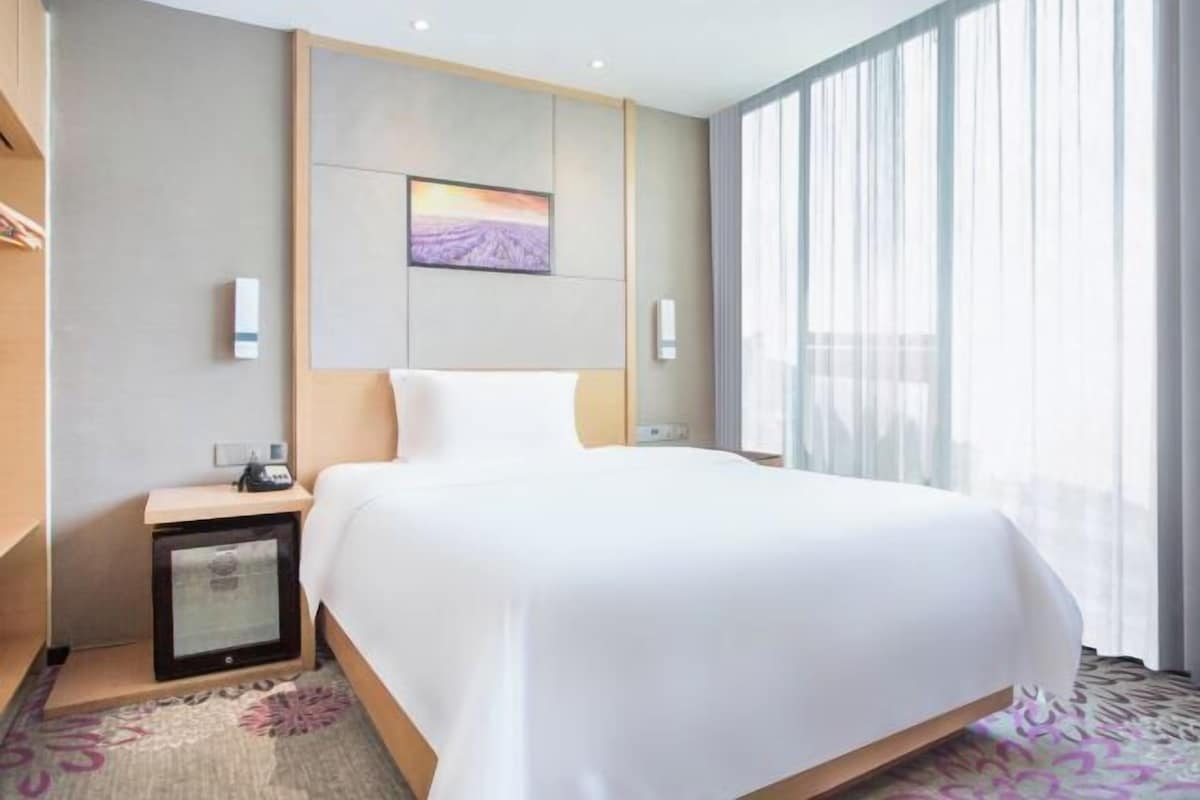 Quarto de hotel com uma cama grande, uma mesa de cabeceira com abajur, um pequeno armário e uma janela ampla com cortinas translúcidas.