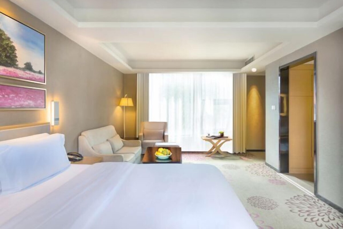 Quarto de hotel com uma cama grande, uma escrivaninha, uma cadeira, um abajur e vista para o exterior pela janela.
