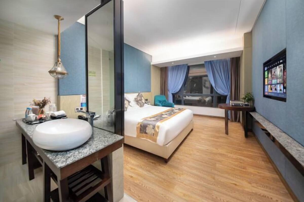 A modern hotel room with a large bed, a round sink, a mirror, a TV, and a window with curtains.